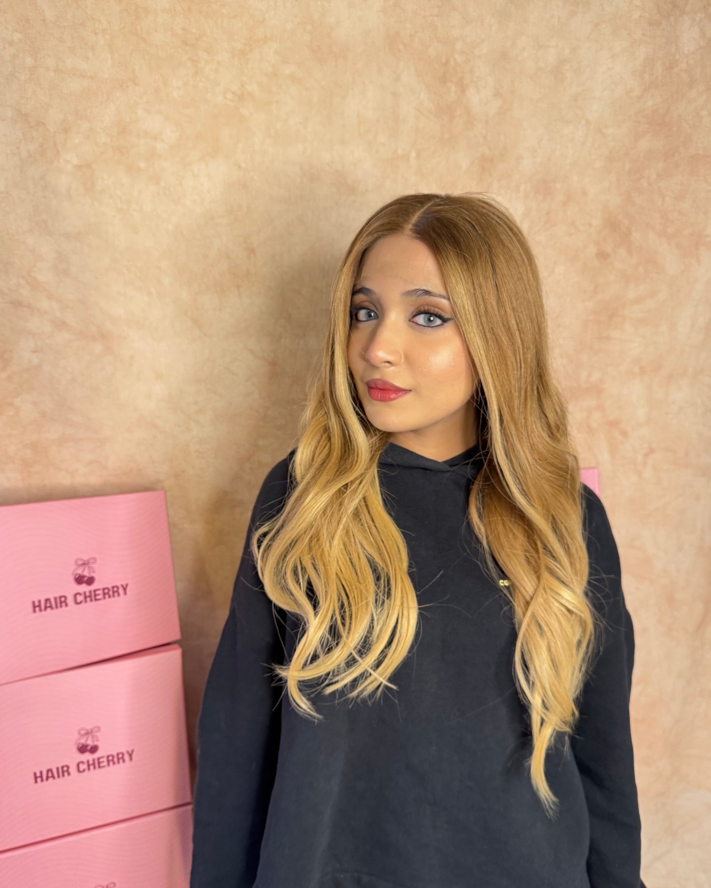 Caramel blonde human hair wig with HD lace and pre-plucked hairline worn by model, showcasing long layered lengths in front of stacked pink HairCherry boxes.